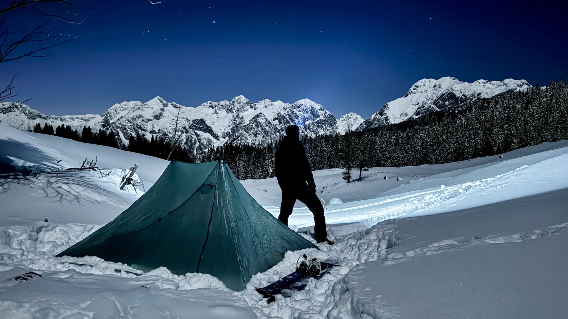 Night photography of a tent pitched in deep snow with snow covered mountains in the background and my silhouette looking at them with my headlamp on